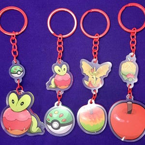 May include: Four keychains with red chains and rings. Each keychain features a different colorful cartoon character design. The designs include a green and red creature, a green and red creature with a ball, a yellow and green winged creature, and a red apple.
