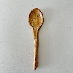 Puede incluir: Una cuchara de cerámica hecha a mano con un esmalte moteado marrón y amarillo. La cuchara tiene una forma única y orgánica.