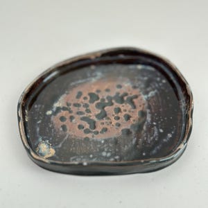 May include: A dark brown ceramic plate with a unique, irregular shape and a speckled, mottled glaze. The glaze has a dark brown base with lighter brown and white speckles.