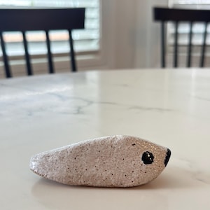 May include: A handmade ceramic sculpture of a small, stylized animal, possibly a seal or a fish. The sculpture is light pink with dark speckles and a black eye. The object is resting on a white marble surface.