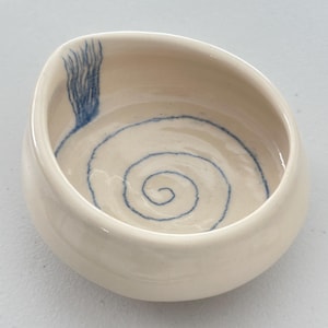 May include: Off-white ceramic bowl with a blue spiral design in the center. A blue brushstroke design is on the side of the bowl. The bowl has a smooth, glossy finish and a rounded shape. The bowl is likely handmade and suitable for serving food.
