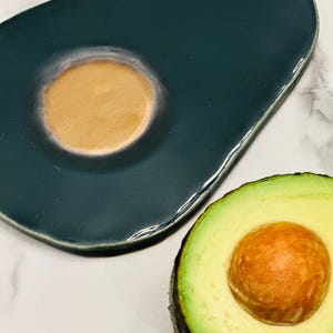 以下が含まれることがあります： アボカドの形をした、茶色の中心部を持つティール色のセラミック製サービング皿と、半分に切られたアボカド。アボカドは緑色の外観と大きな茶色の種があります。この皿は光沢のある仕上がりです。