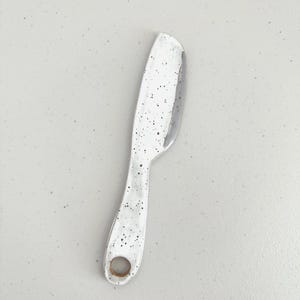 Op de afbeelding: Een wit keramieken botermesje met een zwart gespikkeld patroon over het gehele oppervlak. Het keukengerei heeft een breed, plat blad en een afgerond handvat met een cirkelvormig gat aan de onderkant, afgewerkt met een gladde glans.