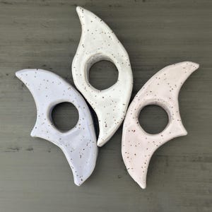 May include: Three ceramic bookmark holders in different colours: white with brown speckles, light purple with brown speckles, and light pink with brown speckles. Each bookmark holder is shaped like a curved leaf with a hole in the centre.