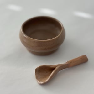 Op de afbeelding: Een kleine bruine keramische kom met een bijpassende bruine keramische lepel. De kom en lepel hebben een gespikkelde textuur.