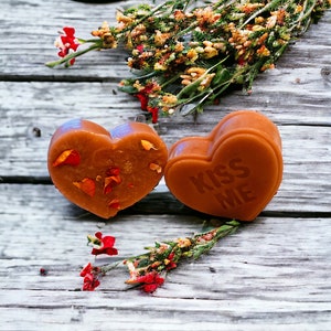 May include: Two heart-shaped soaps on a rustic wooden surface. One soap is decorated with dried flower petals, while the other has the words "Kiss Me" embossed on it.
