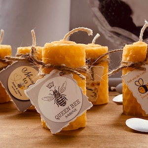 Può includere: Un set di piccole candele gialle con texture a nido d'ape, con spago ed etichette di carta. Le etichette presentano illustrazioni di api e testo che recita "Mean to", "Queen Bee Sabine" e "Suß wie die Liebe Jana & Harry 12.02.21".