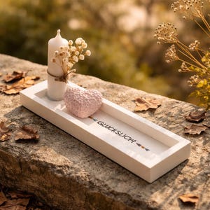 May include: A white rectangular tray with a candle, dried flowers, and a pink heart-shaped object. The tray has the word "GLÜCKSLICH" printed on it. The tray is resting on a stone surface.