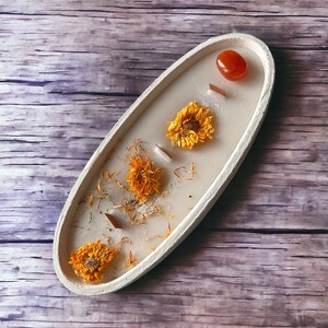 May include: A white oval dish with dried flowers and a small orange stone. The dish is on a wooden surface.