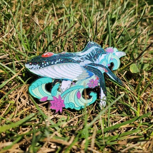 May include: A decorative pin featuring a whale design with a white belly, dark blue body, and accents of teal, pink, and red. The whale is surrounded by stylized waves and lotus flowers, with a glittery finish.