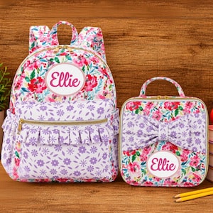 May include: A backpack and a lunch bag set featuring a floral pattern with red and pink flowers on a white background, accented by a purple and white small floral print. Both items have a circular patch with the name Ellie printed in pink.