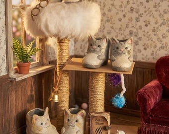 May include: A miniature cat-themed room with a cat tree, cat-shaped planters, and a miniature book titled "Gatito Diary." The cat tree has a fluffy top with two cat figurines wearing glasses. The room is decorated with floral wallpaper.