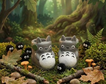 May include: Two gray ceramic planters shaped like the character Totoro, with white bellies and leaf details, sit on a bed of green moss. The planters are surrounded by small black soot sprites, mushrooms, and fallen leaves, creating a whimsical forest scene.