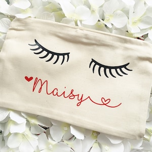 Könnte beinhalten: Eine weiße Kosmetiktasche aus Canvas mit einem roten Herzen und dem Namen "Maisy" in roter Kursivschrift. Die Tasche hat zwei schwarze Wimpern aufgedruckt.