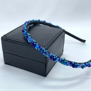 May include: A blue beaded headband with a black elastic band. The headband is on a black box.