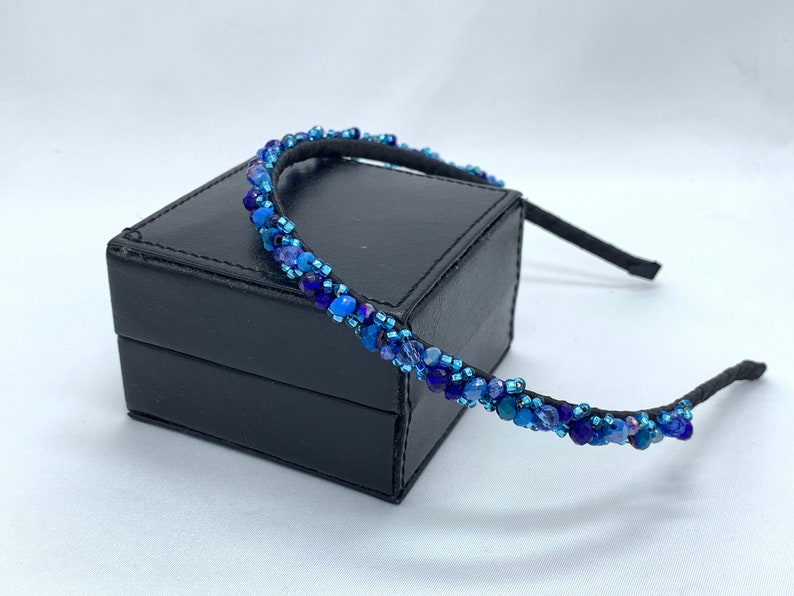May include: A blue beaded headband with a black elastic band. The headband is on a black box.