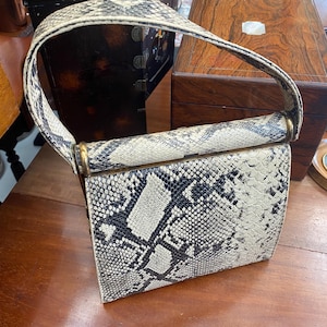 May include: A vintage handbag with a snakeskin pattern in black, white, and gray. The rectangular bag has a curved handle and gold-toned metal accents. The bag is sitting on a wooden surface.