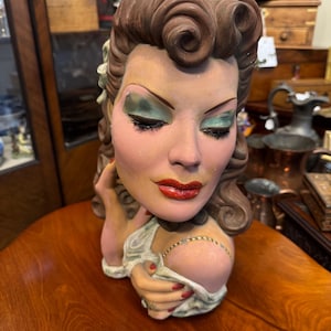 Könnte beinhalten: Vintage Keramikbüste einer Frau mit braunem, gelocktem Haar. Die Büste hat bemalte Gesichtszüge, darunter grüner Lidschatten, roter Lippenstift und eine Halskette. Die Hand der Frau ist in der Nähe ihres Gesichts, und ihre Nägel sind rot lackiert.