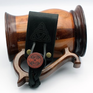 May include: A wooden beer stein with a black leather handle and a Celtic knot design. The handle has a wooden button with a Celtic knot design.