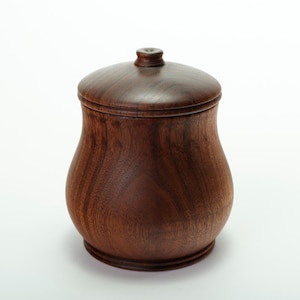 Puede incluir: Un tarro de madera marrón oscuro hecho a mano con tapa. El tarro tiene una forma redondeada y bulbosa con una base ligeramente ensanchada. La tapa es abovedada con un pequeño pomo en la parte superior. La veta de la madera es visible.