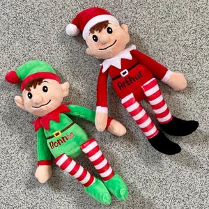 May include: Two plush elf dolls, one in green with "Bonnie" on the front, and the other in red with "Arthur" on the front. Both dolls have striped legs, elf hats, and black shoes. The dolls are lying on a gray surface.