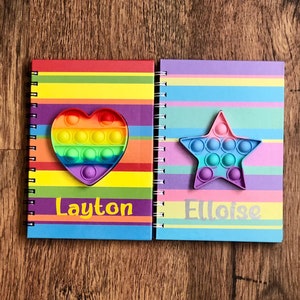 May include: Two spiral-bound notebooks with rainbow striped covers. One has a heart-shaped pop-it toy, the other a star-shaped pop-it toy. The name "Layton" is printed on the first notebook, and "Eloise" on the second.