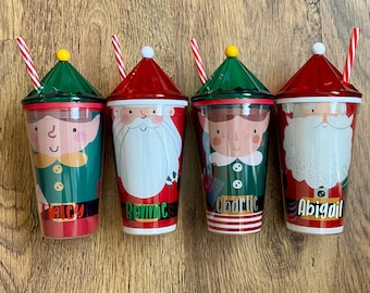 May include: Four Christmas-themed plastic cups with lids and straws. Each cup features a different character: an elf named Stacy, Santa Claus named Bernie, an elf named Charlie, and Santa Claus named Abigail. The cups have red and green accents.