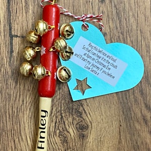May include: A red wooden Christmas bell with gold bells and snowflake accents. The bell has a wooden handle with the name "Finley" printed on it. A blue heart-shaped tag with text is attached.