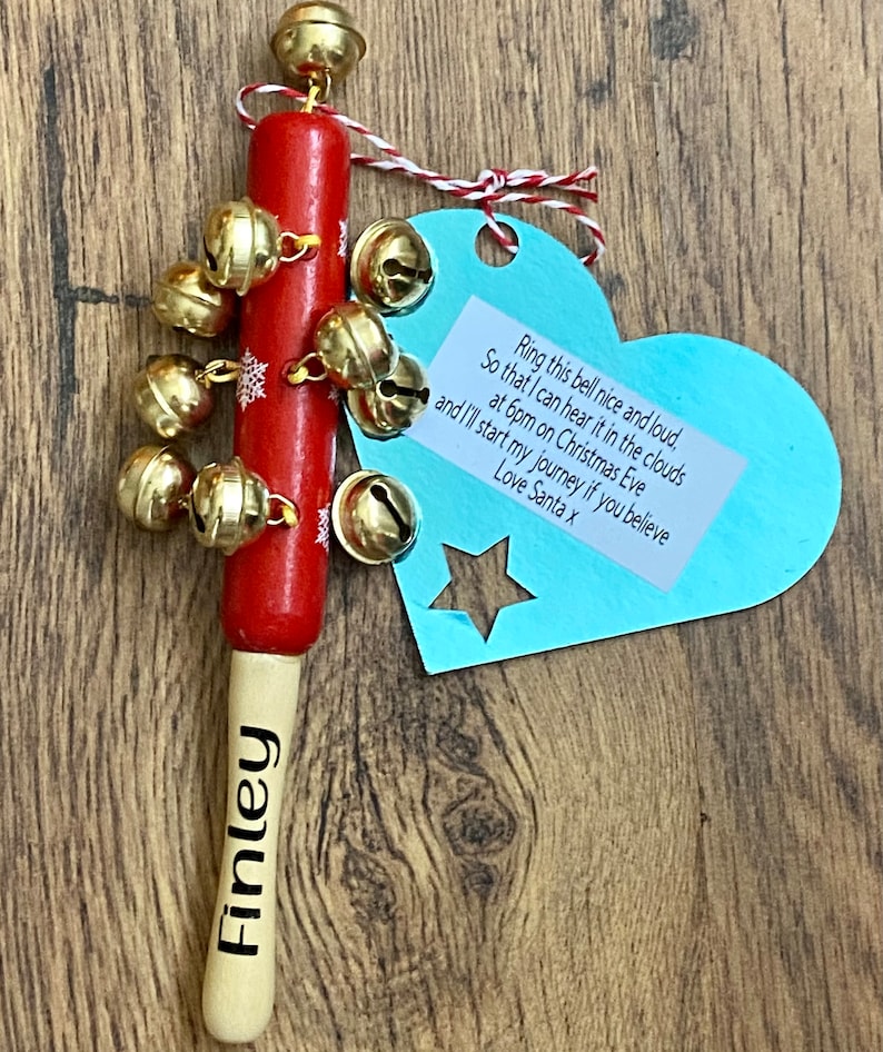 May include: A red wooden Christmas bell with gold bells and snowflake accents. The bell has a wooden handle with the name "Finley" printed on it. A blue heart-shaped tag with text is attached.