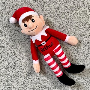 May include: A plush elf doll in a red outfit with white and red striped legs and black shoes. The elf has a red hat with a white trim, brown hair, and the name "Arthur" is printed on the chest.
