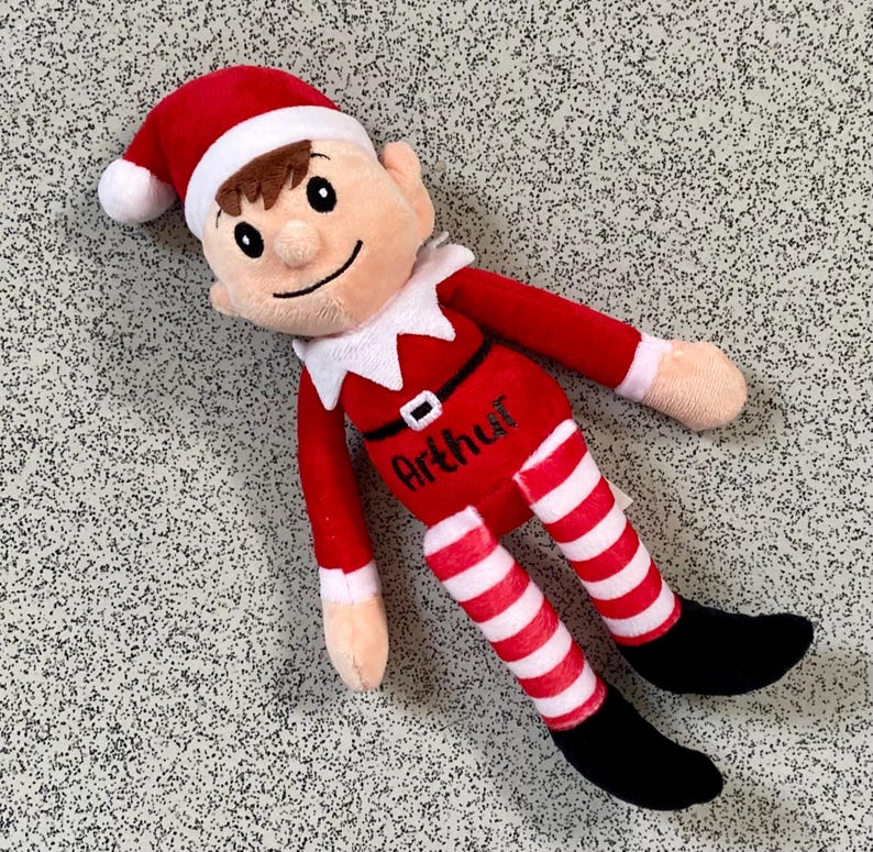 May include: A plush elf doll in a red outfit with white and red striped legs and black shoes. The elf has a red hat with a white trim, brown hair, and the name "Arthur" is printed on the chest.