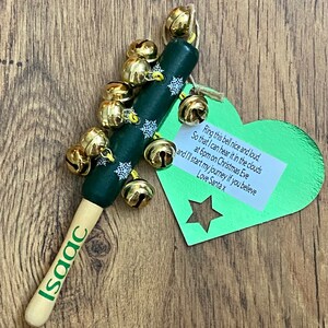 May include: A Christmas-themed wooden bell stick with gold bells and snowflake accents. The handle is painted green with the name "Isaac" in green. A green heart-shaped tag with text is attached.