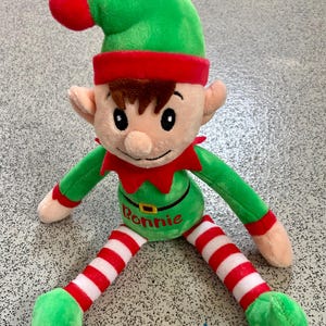 May include: A plush elf doll with a green hat, red trim, and a red pom-pom. The elf has a green body, red and white striped legs, and green feet. The name "Bonnie" is embroidered on the belly. The doll is designed for holiday decor.