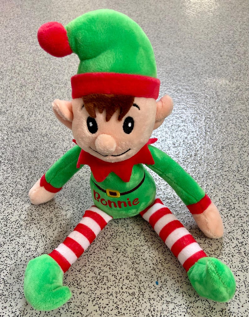 May include: A plush elf doll with a green hat, red trim, and a red pom-pom. The elf has a green body, red and white striped legs, and green feet. The name "Bonnie" is embroidered on the belly. The doll is designed for holiday decor.