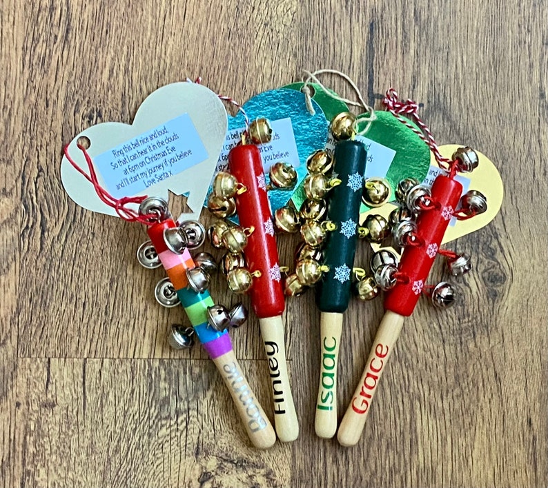 May include: Four wooden Christmas bell ornaments. The bells are attached to painted wooden sticks in red, green, and rainbow colors. Each stick has a name written on it: Fintey, Isaac, and Grace. The ornaments have small tags with text.