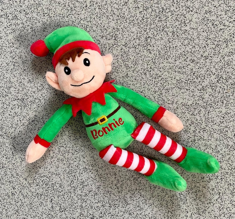 May include: A plush elf doll with a green outfit, red collar, and a matching hat with a red pom-pom. The elf's legs are striped red and white. The name "Ronnie" is embroidered on the doll's torso.