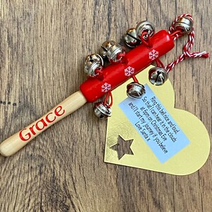 May include: A red wooden Christmas bell with silver bells attached. The handle is light wood with the name "Grace" in red. A gold tag with a star cutout and text is attached.