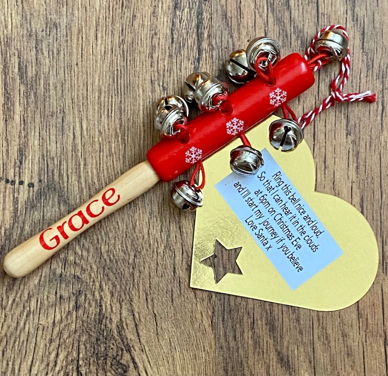 May include: A red wooden Christmas bell with silver bells attached. The handle is light wood with the name "Grace" in red. A gold tag with a star cutout and text is attached.