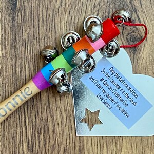 May include: A wooden tambourine with a rainbow-colored handle and silver bells. The handle is inscribed with the name "Bonnie". A heart-shaped tag with text is attached, including the phrase "Ring this bell nice and loud".