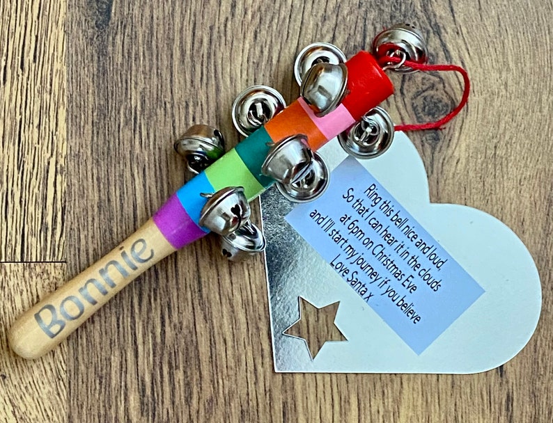May include: A wooden tambourine with a rainbow-colored handle and silver bells. The handle is inscribed with the name "Bonnie". A heart-shaped tag with text is attached, including the phrase "Ring this bell nice and loud".