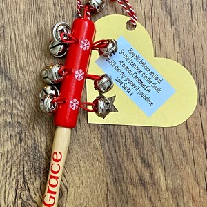 May include: A red wooden Christmas bell with silver bells and snowflake accents. The handle is natural wood with the name "Grace" in red. A yellow heart-shaped tag with text is attached with red and white twine.