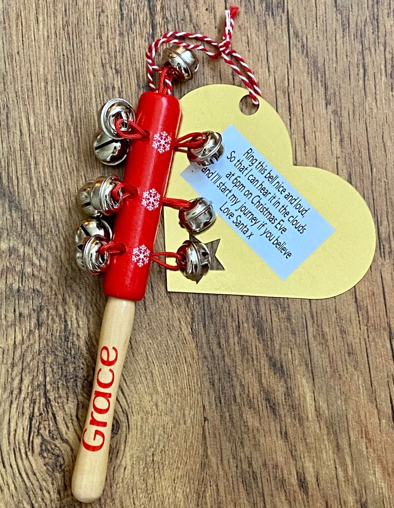 May include: A red wooden Christmas bell with silver bells and snowflake accents. The handle is natural wood with the name "Grace" in red. A yellow heart-shaped tag with text is attached with red and white twine.