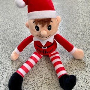May include: A plush elf doll with a red hat and outfit, white collar, and striped red and white legs. The doll has a smiling face, black shoes, and the name "Arthur" on its belly. A festive holiday decoration.