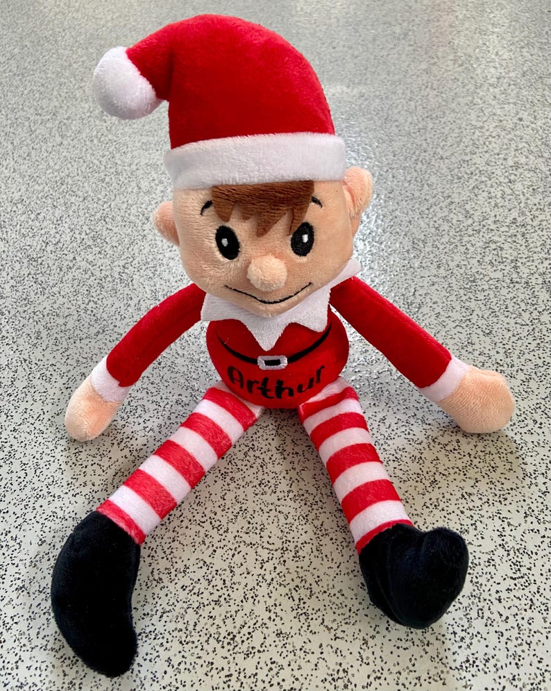 May include: A plush elf doll with a red hat and outfit, white collar, and striped red and white legs. The doll has a smiling face, black shoes, and the name "Arthur" on its belly. A festive holiday decoration.
