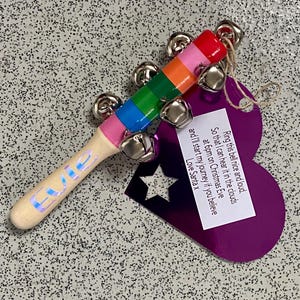 May include: A wooden Christmas bell with a rainbow-colored handle and silver bells. The handle has the name "Evie" in iridescent lettering. A purple tag with a star cutout and a message from Santa is attached.