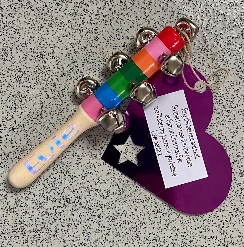 May include: A wooden Christmas bell with a rainbow-colored handle and silver bells. The handle has the name "Evie" in iridescent lettering. A purple tag with a star cutout and a message from Santa is attached.