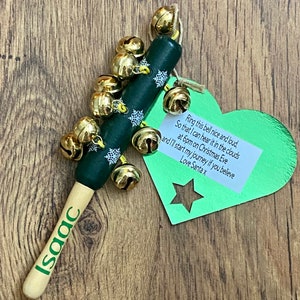 May include: A Christmas-themed wooden bell stick with the name "Isaac" printed on the handle. The dark green body is adorned with white snowflakes and gold bells. A green heart-shaped tag with text is attached.