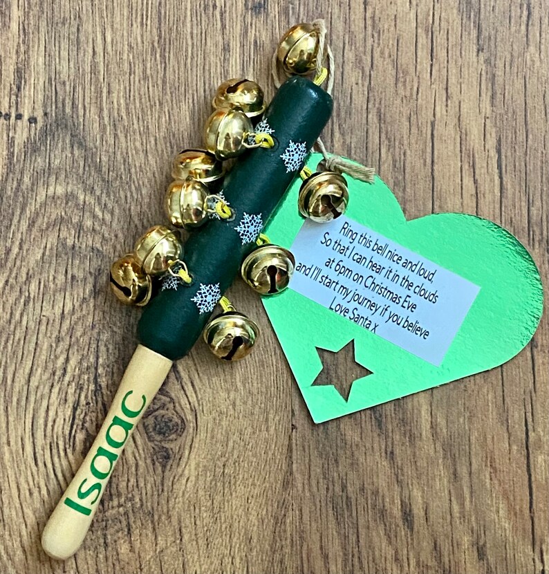 May include: A Christmas-themed wooden bell stick with the name "Isaac" printed on the handle. The dark green body is adorned with white snowflakes and gold bells. A green heart-shaped tag with text is attached.