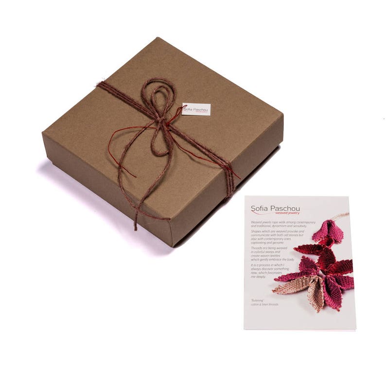 May include: A brown cardboard box with a red and brown twine bow. The box is tied with a red ribbon and has a small white tag with the text "Sofia Paschou" on it. The box is sitting on a white surface.