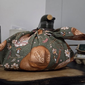 Pode incluir: Um saco de pão verde com um padrão de pães e elementos florais. O saco de tecido é amarrado no topo, cobrindo um eletrodoméstico de cozinha. O design inclui ilustrações de pão, flores, abelhas e ferramentas de jardinagem.
